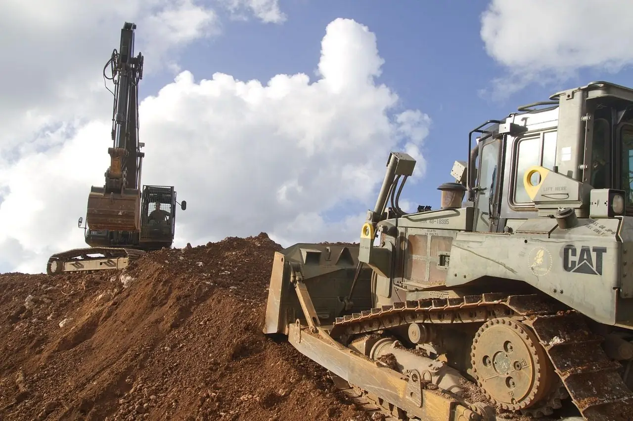 Yellow bulldozer in front of an excavator on a dirt site under a cloudy sky